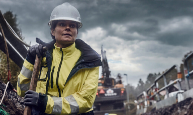 Anleggsarbeider i gul refleksjakke med spade foran gravemaskin på byggeplass under grå himmel.