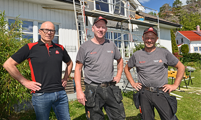 Tre håndverkere i grå og svart uniform foran hvitt hus med stillas og verktøy i hagen.