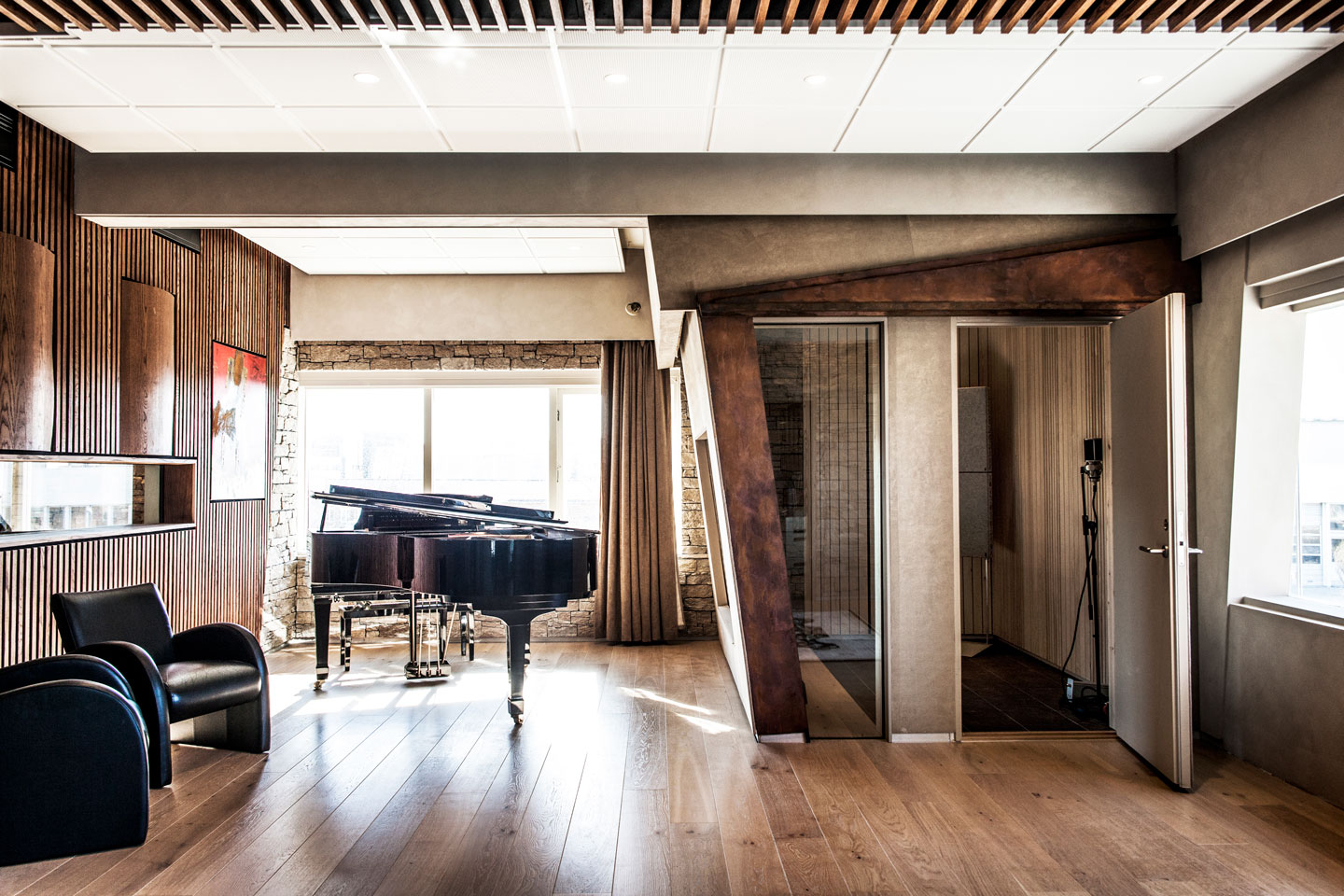Moderne musikkstudio med svart flygel på lyst eikegulv, mørke trepaneler, lærstoler og vokalbås