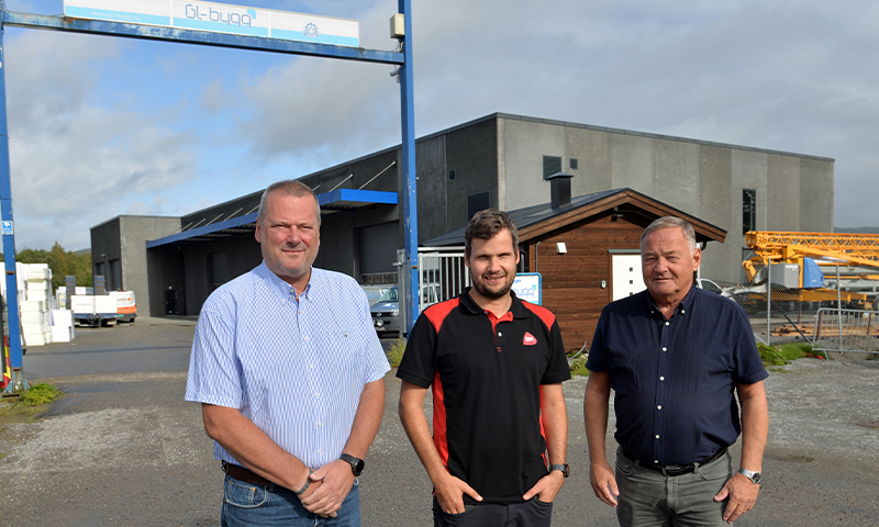 Tre menn på grusplan foran et grått fabrikkbygg med blå portal og en gul kran i bakgrunnen.