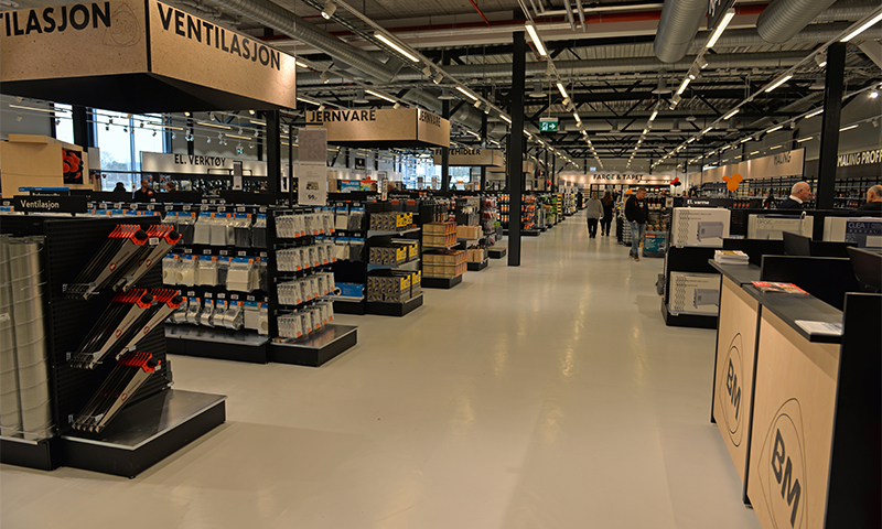 Lyst moderne byggevarehus med hvit gulvflate, svarte hyller, ‘Ventilasjon’-skilt og rør i tak.