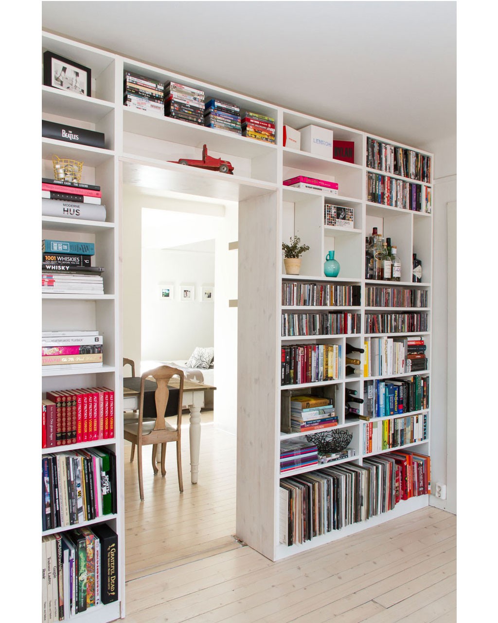 Hvit bokhylle rundt døråpning med fargerike bøker, plater, DVD, rød lekebil og turkis vase.