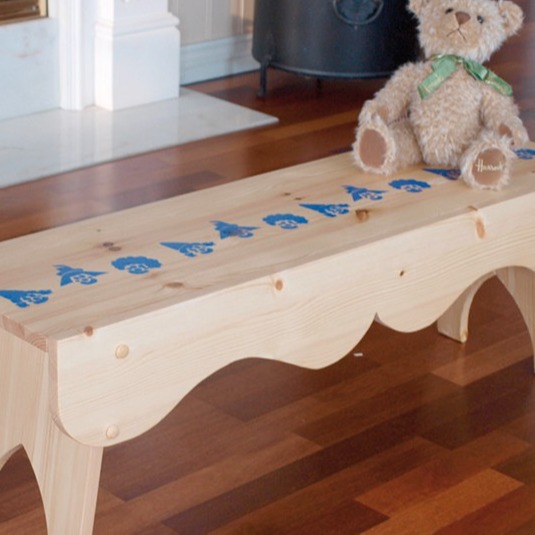 Lys furubenk med blå malte figurer og bamse med grønt bånd på brun parkettgulv foran peis.