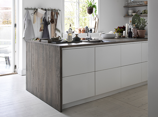 Lys skandinavisk kjøkkenøy i hvitt og lyst tre, med hengeutstyr og grønne planter ved vindu.