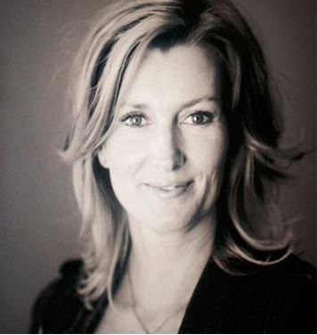 Person med skulderlangt lyst hår og mørk jakke foran grå bakgrunn.