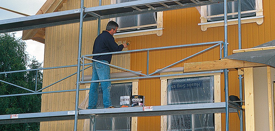 Mann maler gul trekledning på husfasade fra stillas under vindu.