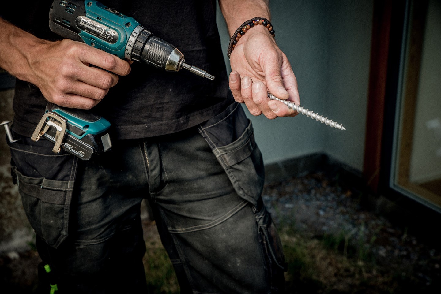 Mann i sort arbeidstøy holder en grønn-svart drill og en stor sølvskrue i hver hånd
