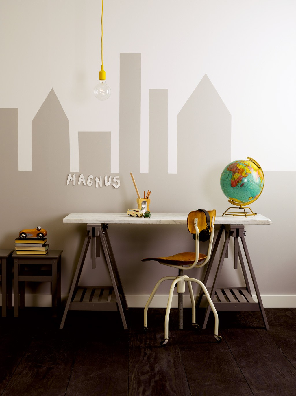 Barneskrivebord mot grå bysilhuett med “Magnus”, hvit bordplate, stol, globus og gul lyspære.