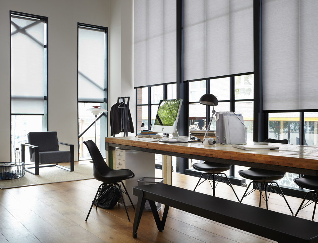 Moderne kontor med lang trebenk, svarte stoler, PC, gulv-til-tak-vinduer med lyse rullegardiner.