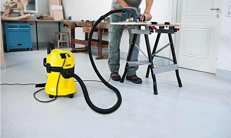 Person sager plank på grå arbeidsbenk i lyst verksted, tilknyttet gul støvsuger.