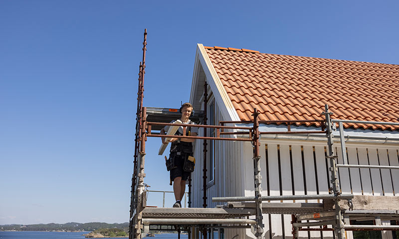 Arbeider på stillas ved hvitt hus med oransje takstein under klar blå himmel