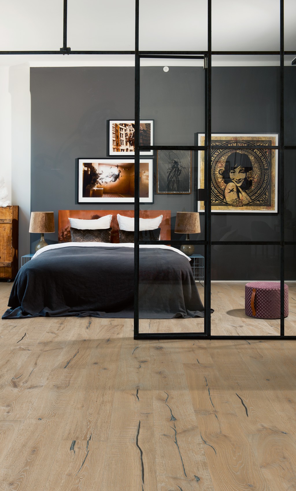 Lyst tregulv, svart glassvegg med rammer, dobbeltseng med grått sengeteppe og veggkunst.