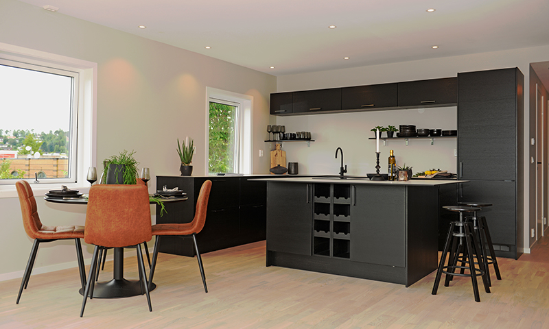 Åpent lyst kjøkken med sort kjøkkenøy, barstoler, rundt spisebord med brune stoler og vinduer.