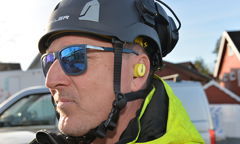 Byggearbeider med svart hjelm, neon gul jakke og gult hørselsvern ved anleggsområde.
