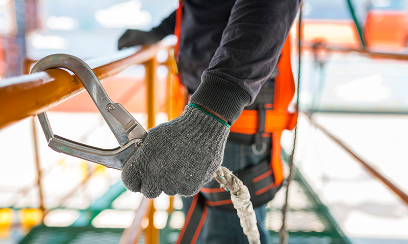Nærbilde av hender i grå hansker som fester oransje sikkerhetskrok til stillasrekkverk