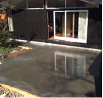 Nylagt grå betongflate foran hus med sort trepanel, hvit skyvedør og speilende overflate.