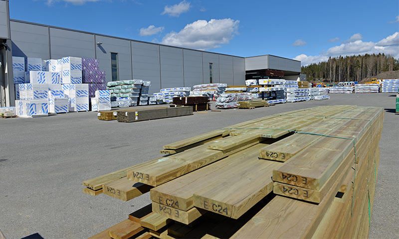 Åpent anlegg med stabler av grønne treplanker og hvite byggepakker ved grå lagerhall