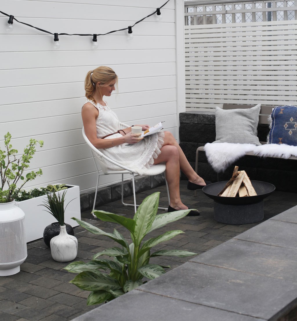 Kvinne i hvit kjole leser og drikker kaffe på grå-hvit uteplass med planter og bålfat