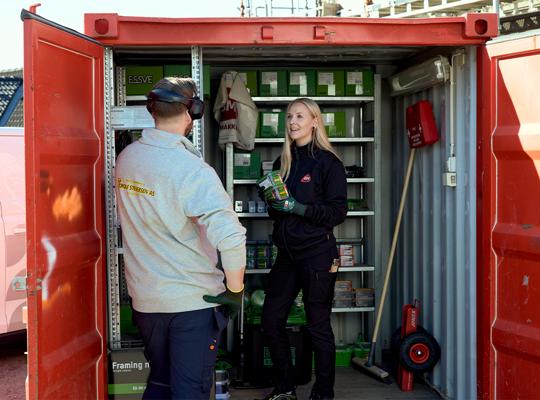 Rød container med hyller av grønne esker, to arbeidere i arbeidstøy med vernehansker