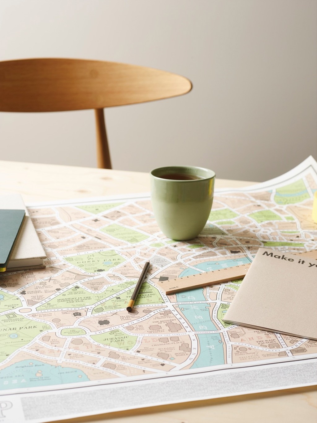 Utsnitt av pastellkart på trebord med grønn tekopp, blyant og notatbok foran trestol.