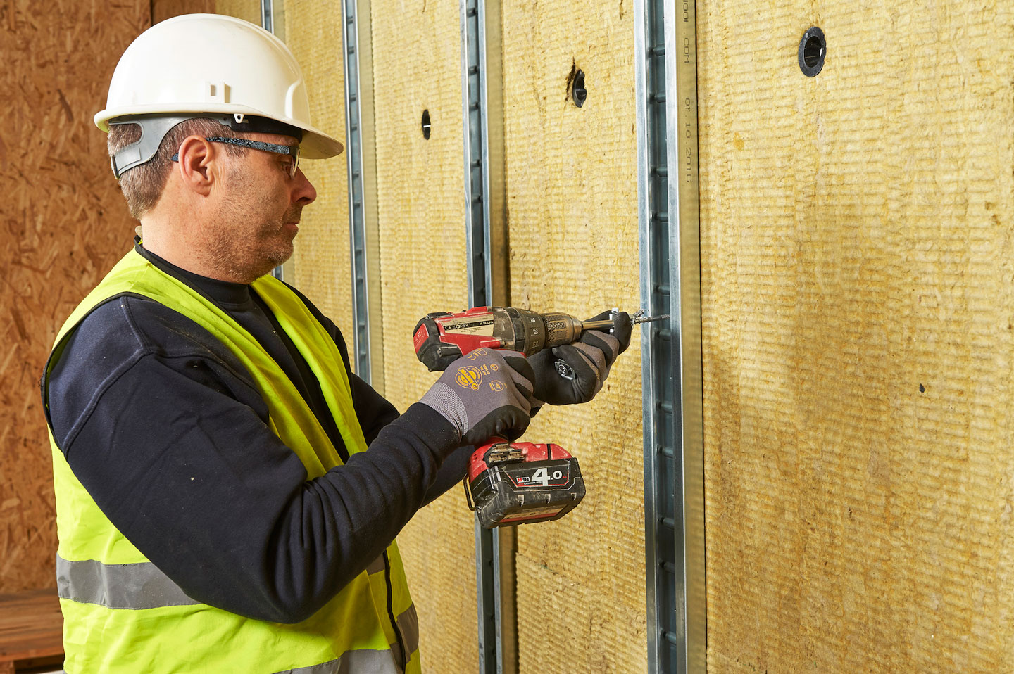 Håndverker i hvit hjelm og gul vest bruker drill på metallstender mot gul isolasjon
