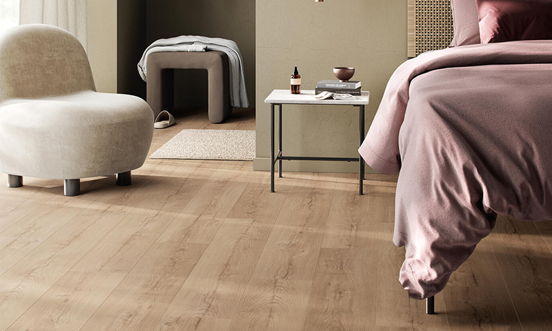 Soverom med dus rosa seng, lyst tregulv, beige lenestol og hvitt nattbord med bok og flaske.