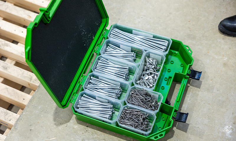 Grønn verktøykasse med åpnet lokk, plastbokser med sorterte stålskruer på betonggulv.