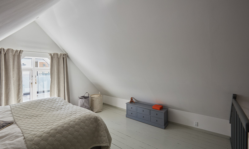 Lyst loftsoverom med skråtak, seng ved vindu med beige gardiner, kurver og blå kommode med rød bok.