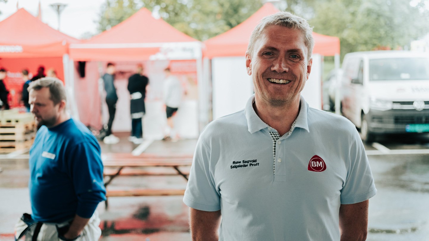 Mann i lys blå BM-polo foran røde messetelt, våte benker og hvit varebil.