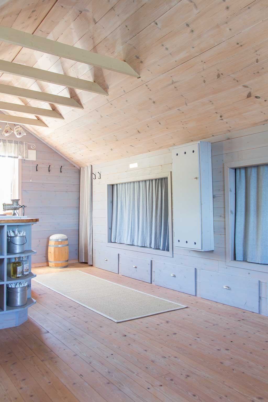 Lyst treloft med skråtak, barhylle med flasker, tønne, løper og sittebenk med gardiner.