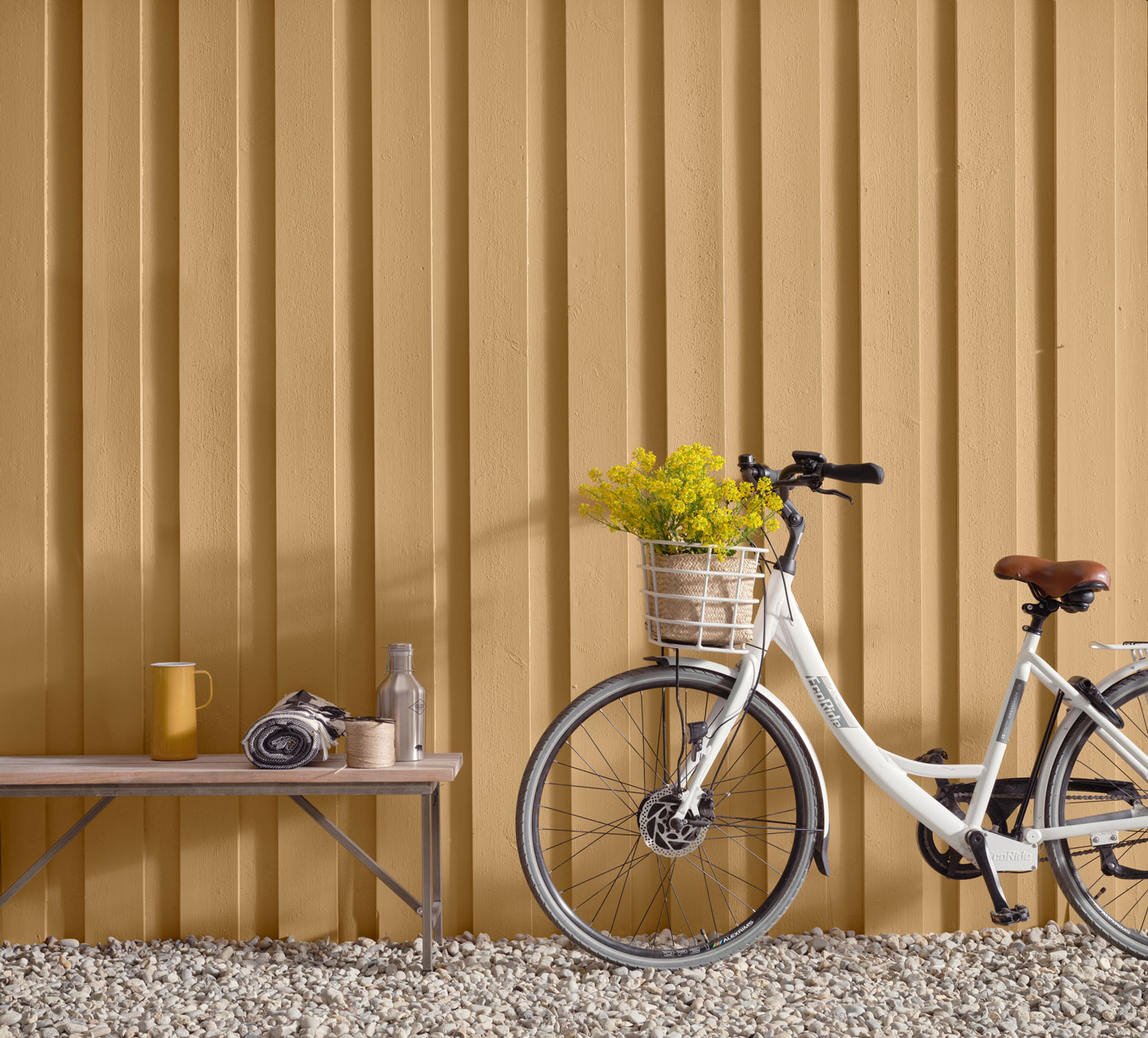 Hvit sykkel med blomsterkurv ved gul vegg, benk med kopp, teppe og flaske på grus.