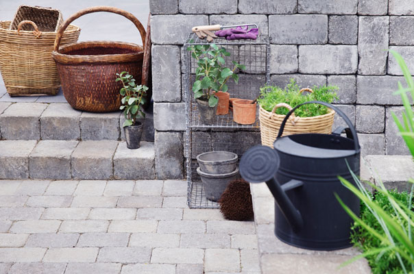 Grå steinplatting med flettekurver, pottede grønne planter på trådhylle og svart vannkanne.
