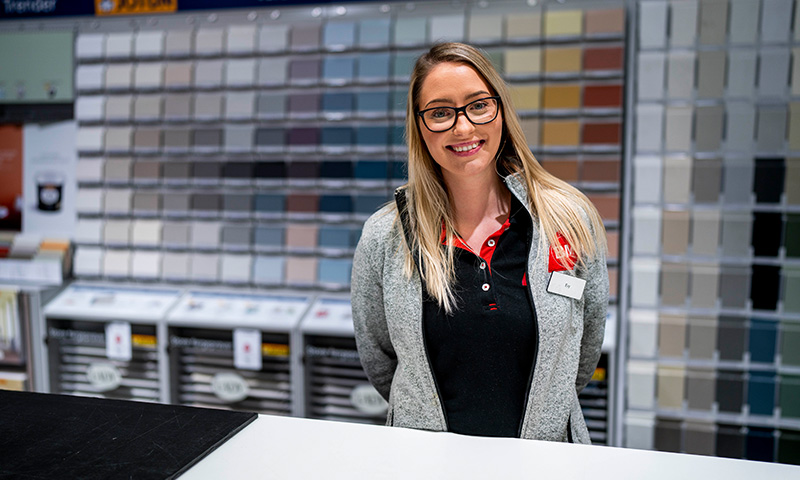 Ansatt i grå genser bak disken i malingsbutikk med fargekart i bakgrunnen