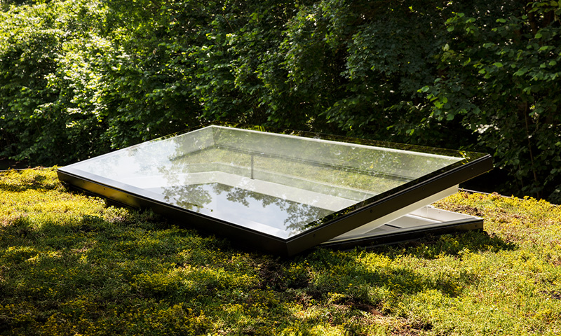 Åpen rektangulær takluke med sort ramme på grønn mosegrodd takflate, foran tett skog