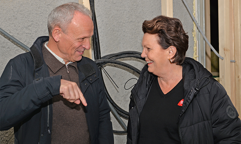 To personer i mørke jakker står ved en byggeplass med kabler i bakgrunnen, én peker.