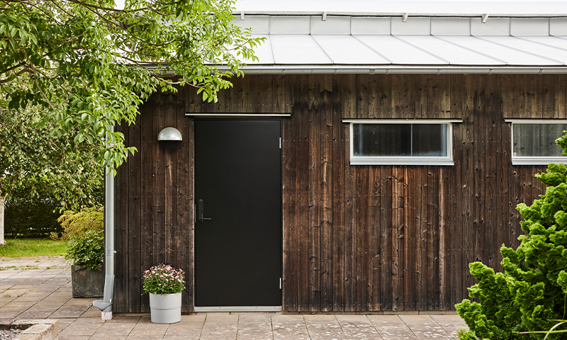 Brun tømmerhytte med svart dør, to smale vinduer og potte med rosa blomster i hage.