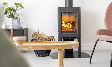 Interiør med svart vedovn med flammer, vedkurv, lys trebord, grønn plante og rosa stol