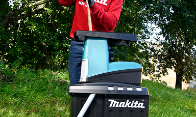 Person i rødt betjener en turkis Makita-batterilader på svart Makita-kasse i gress.