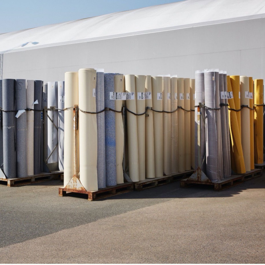 Store ruller med gulvbelegg i beige, grått, blått og gult på paller utenfor hvit bygning.