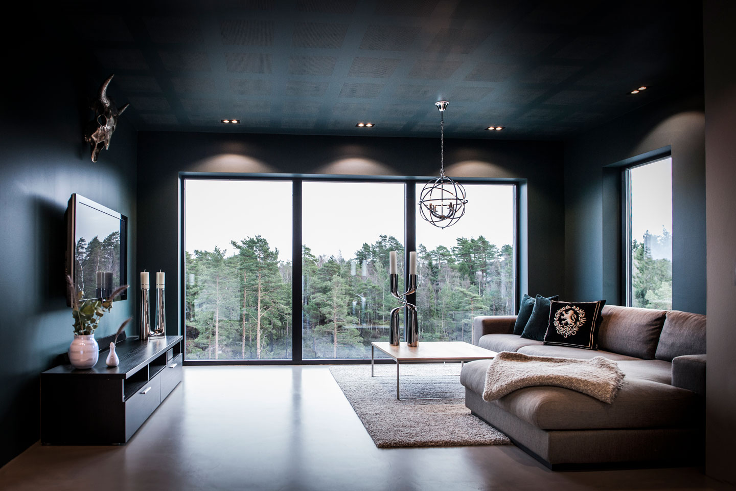 Moderne mørk stue med panoramavindu mot skog, grå sofa med puter, metalllysestaker og globelys.