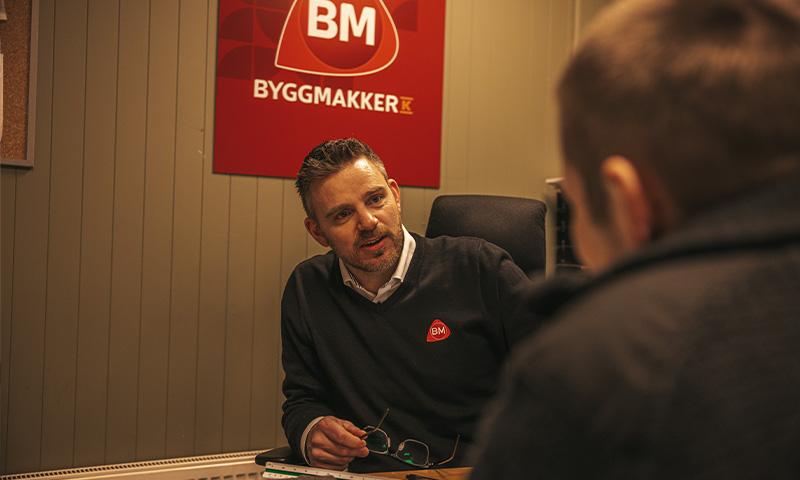 Byggmakker-ansatt i mørk genser ved skrivebord, rødt logo-skilt på grå vegg, snakker med kunde.
