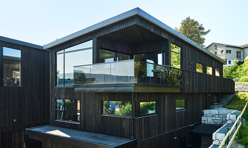 Moderne svart trehus med store vinduer og glassbalkong på grønn skråning