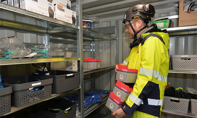 Arbeider i gul sikkerhetsjakke og hjelm i lager med metallhyller fulle av verneutstyr.