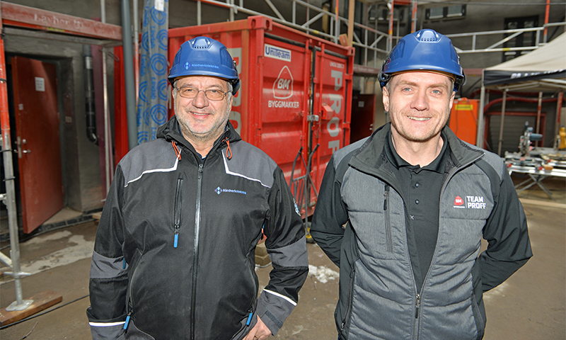 To menn i blå vernehjelmer og mørke jakker foran rød container på byggeplass.