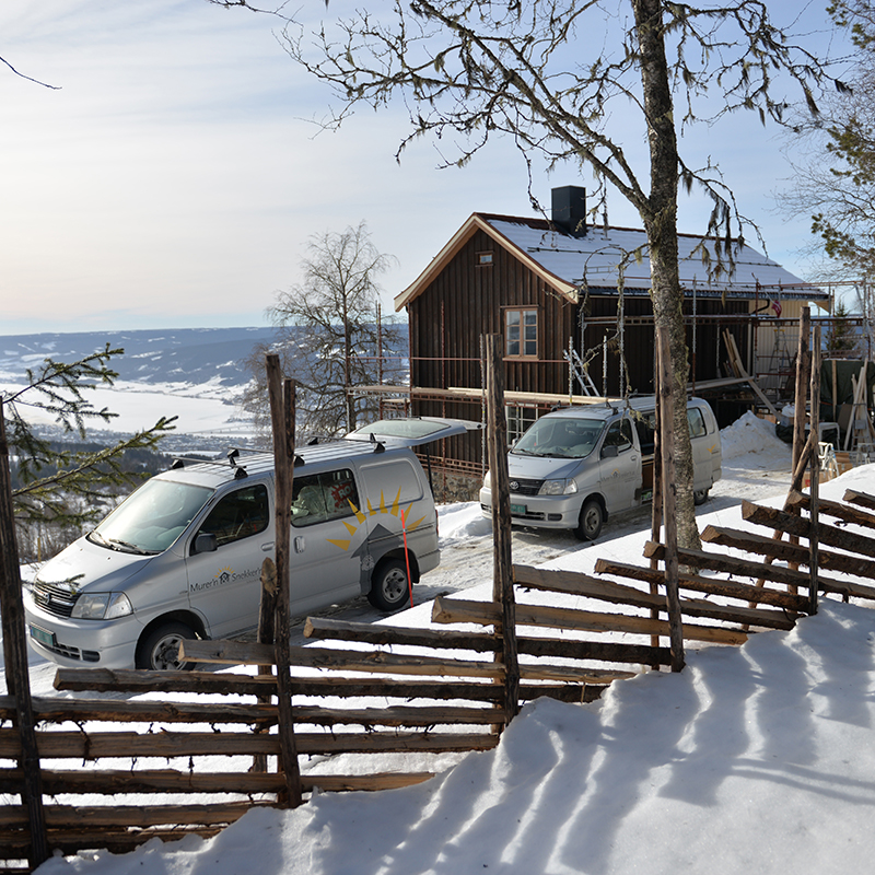To sølvgrå varebiler utenfor brun hytte under oppussing i snødekt fjellandskap med blå himmel.