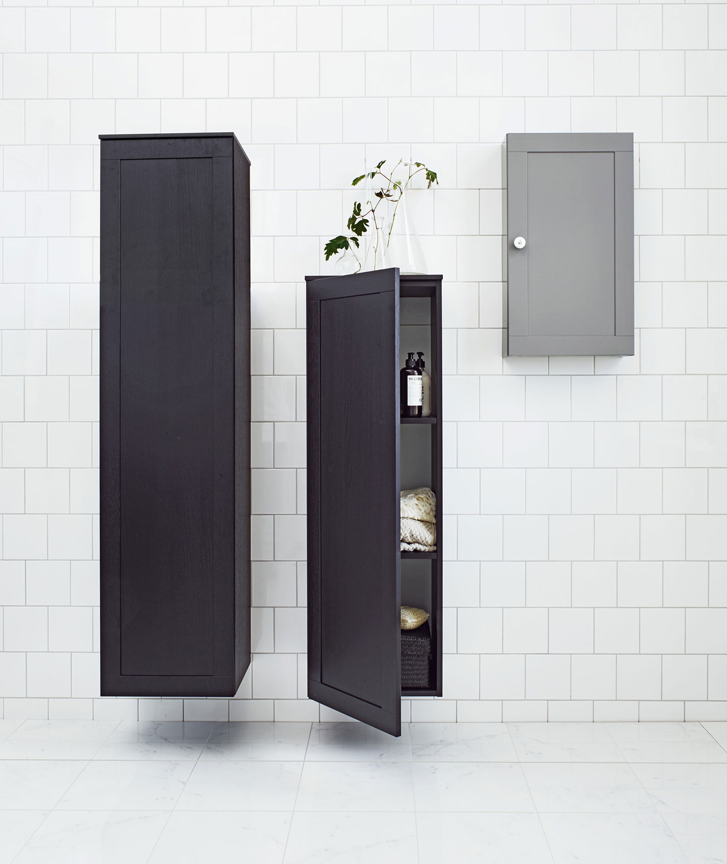 Hvit flisvegg med to svarte vegghengte skap (ett åpent med håndklær og flasker), grå skap med plante