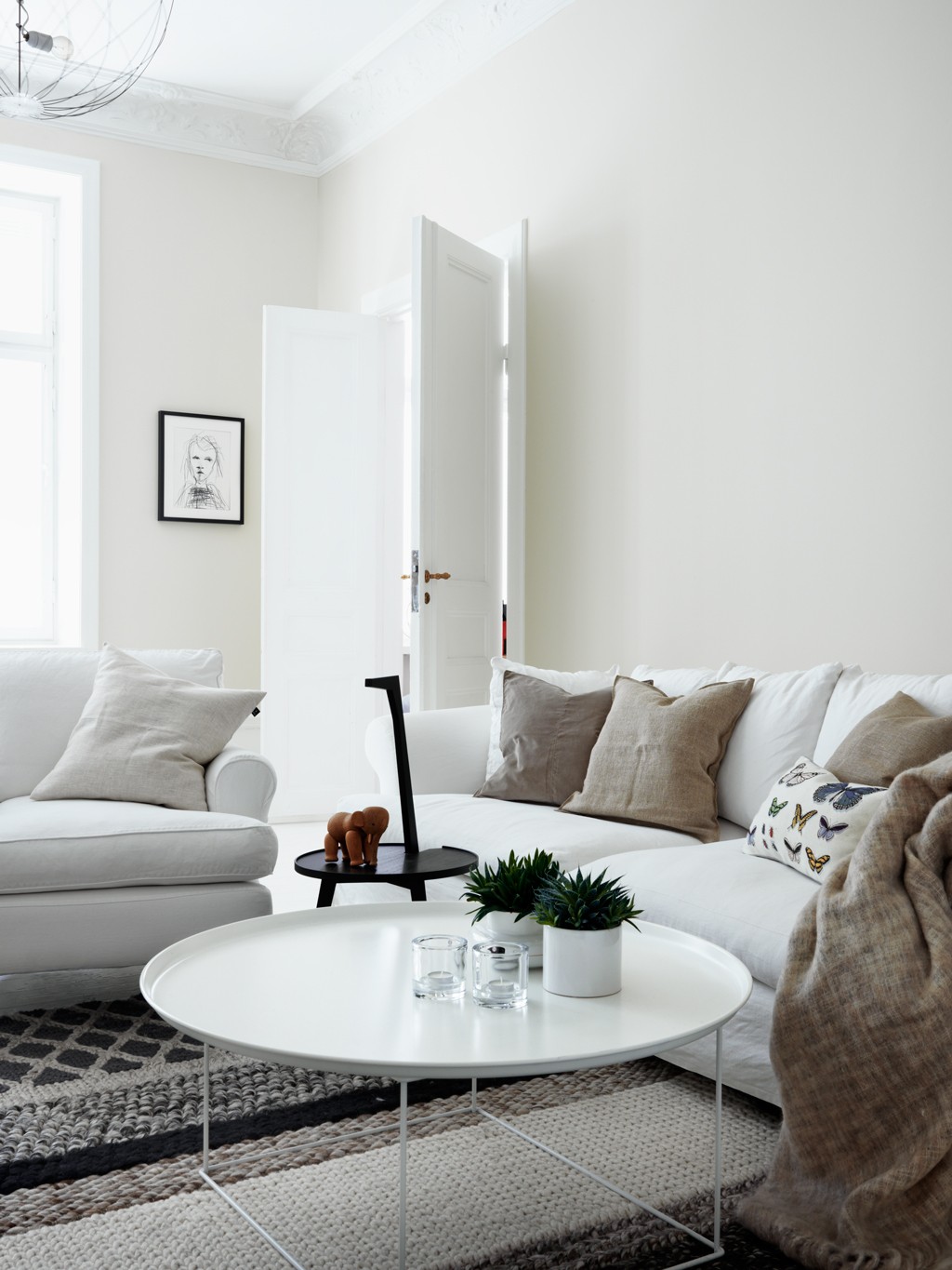 Lyst skandinavisk stue med hvit sofa, beige puter, rundt hvitt bord, grått teppe og grønne planter.