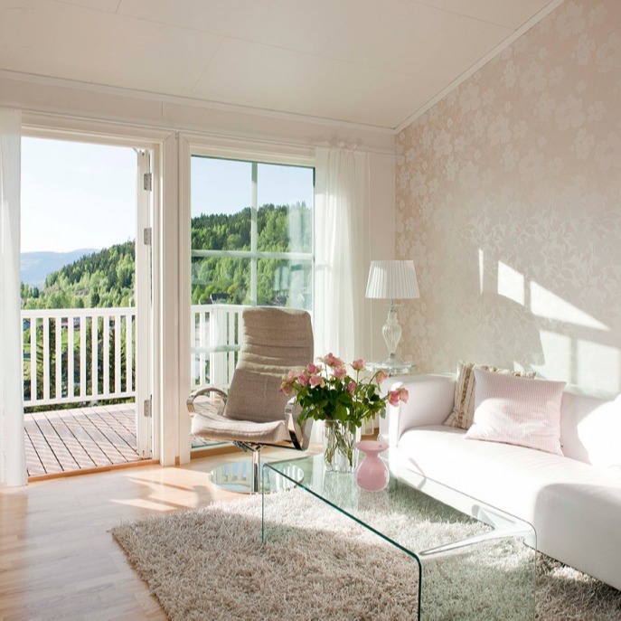 Lyst stue med hvit sofa, glassbord med rosa blomster, lenestol ved åpen balkongdør mot grønne åser.