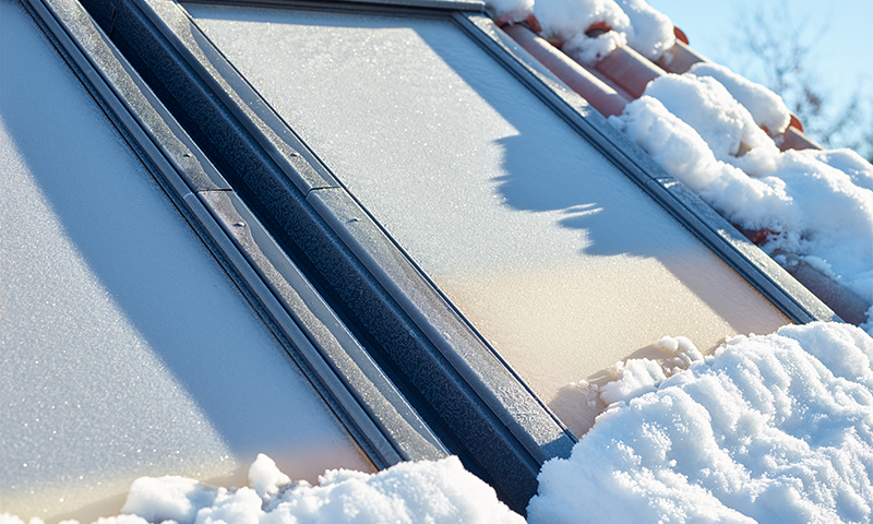 To solcellepaneler på rødt tegltak dekket av snø og rimfrost i solskinn.