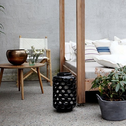 Ute-lounge med himmelsseng, hvite puter, trebord med bronsebolle, svart lanterne og grønn plante.